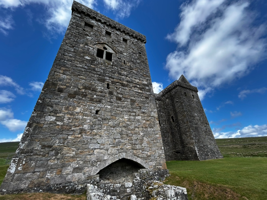 Two tall, stone towers of a ruined castle, set against a bright blue sky with fluffy white clouds. The towers are made of grey stone, showing the effects of age and weathering. The foreground shows grass and the base of one of the towers. The overall impression is one of age, history, and a somewhat desolate beauty.