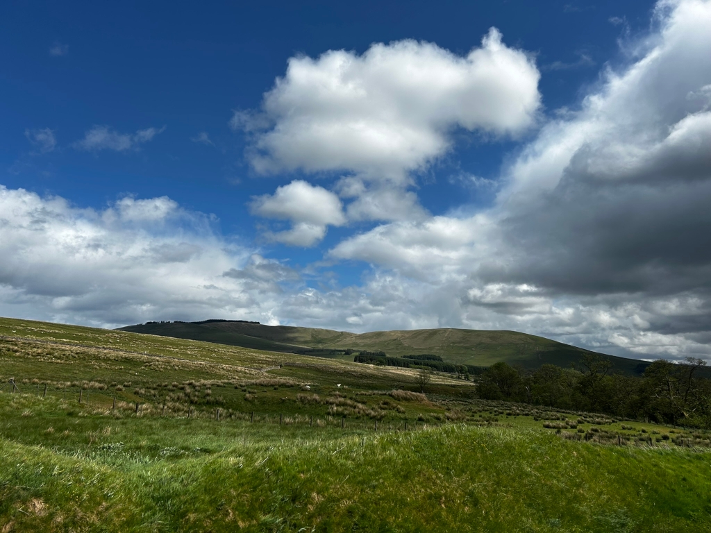 Picturesque landscape under a partly cloudy sky. The foreground is dominated by lush green grass, gently sloping towards a distant hill or low mountain. The hill is covered in vegetation, appearing brown and green. The sky is a vibrant blue, punctuated by fluffy white clouds that gradually transition into darker, more dramatic clouds towards the right. The overall scene evokes a feeling of peaceful, open countryside.