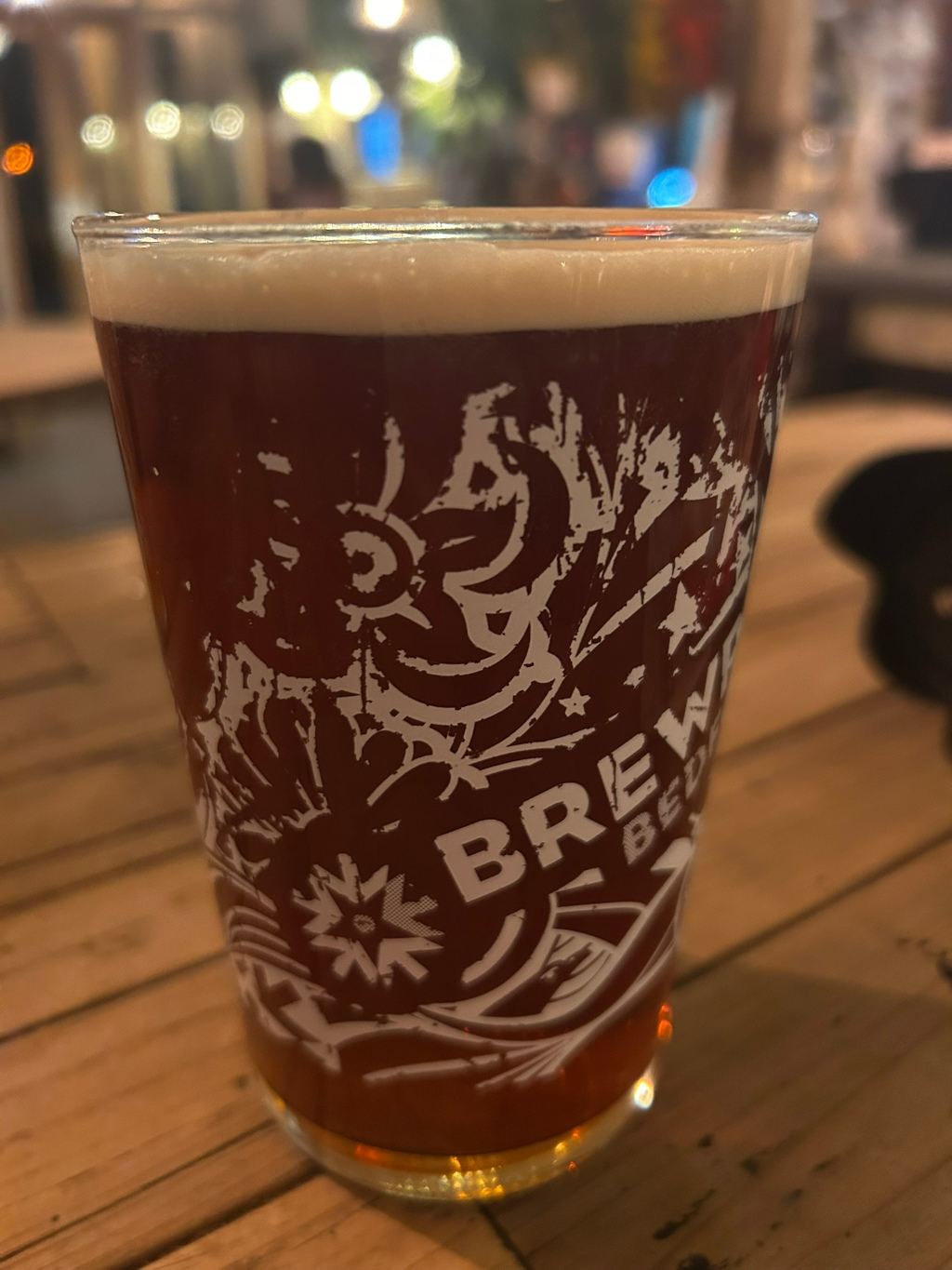 Pint glass of dark beer on a wooden table. The glass has a design printed on it, featuring stylised graphics and the word BREW partially visible. The background is blurred, suggesting a pub or bar setting. The focus is primarily on the beer glass.