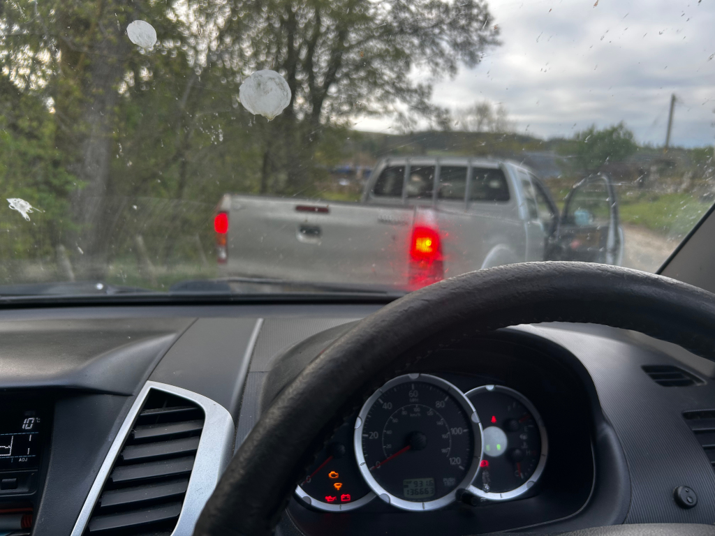 View from inside a car, looking through a somewhat dirty windshield at another vehicle ahead. The car in the foreground appears to be a pickup truck, and a similar model pickup truck is visible ahead, partially obscured. The background shows a rural or country setting with some trees and overcast sky.