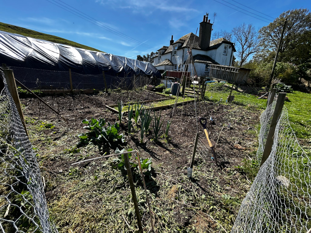 Vegetable garden in the process of being prepared for planting. It's a partially tilled plot of land, enclosed by chicken wire fencing, with some young plants already visible (leeks and possibly cabbages). Gardening tools are lying on the ground. In the background a stone house is visible. The overall impression is of springtime preparation for gardening, located in a rural setting.