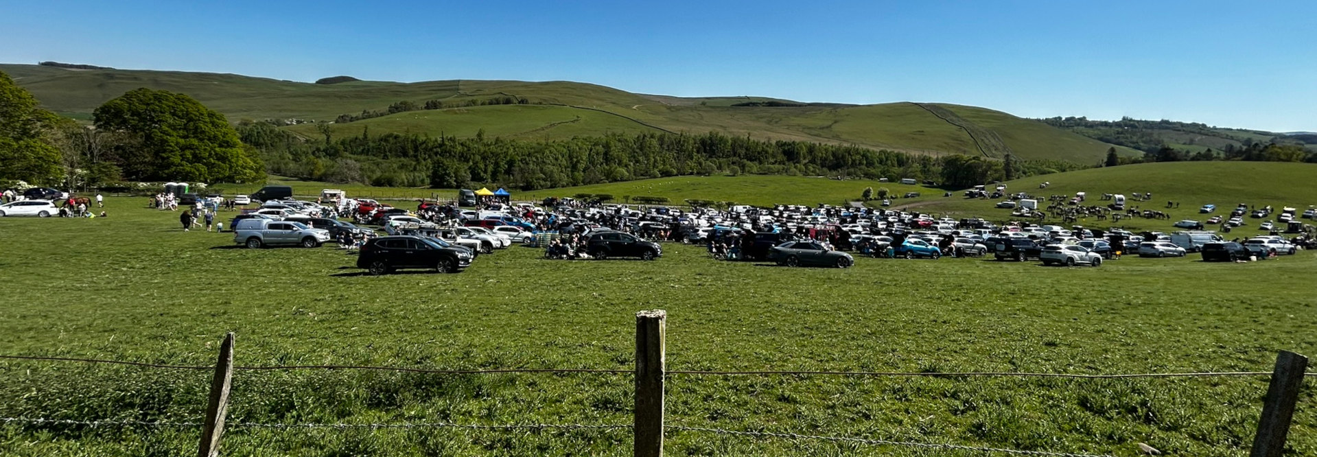Large number of cars parked on a grassy field. Many people are visible amongst the vehicles, suggesting an outdoor event or gathering. The background reveals rolling green hills under a clear blue sky. A wire fence is in the foreground.