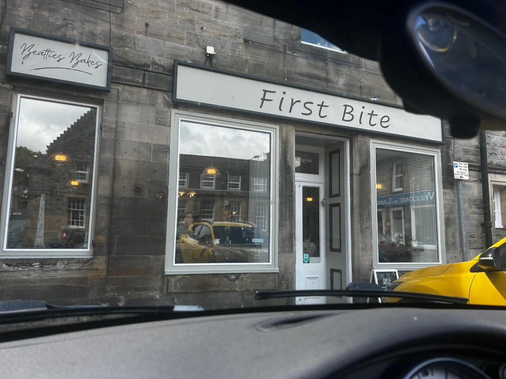 Storefront called First Bite, located next to a business called Beatties Bakes. The view is from inside a car, looking out at the bakery. The windows reflect the street and buildings opposite, and there are people visible inside First Bite. A yellow car is partially visible parked near the storefront. The overall impression is of a quaint, possibly British, bakery storefront.