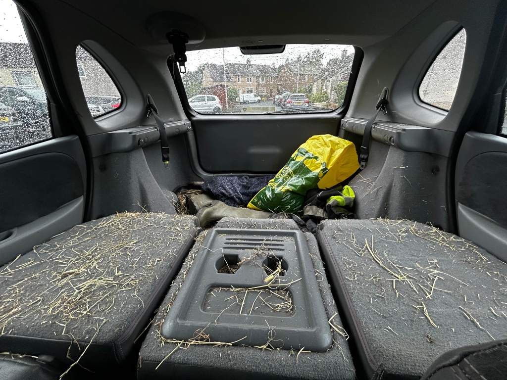 Back of Charlie's car, specifically the rear seats and cargo area. The seats and floor are covered in a significant amount of hay or straw. A yellow bag, possibly containing feed or fertilizer, is visible in the cargo area along with other items, which appear to be some kind of work boots and a hi-vis jacket. The car appears to be used for transporting materials related to agricultural or landscaping work. The overall impression is one of a vehicle used for practical, possibly rural, purposes.