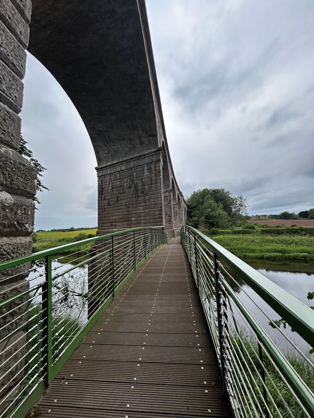 Modern pedestrian bridge extending from the viewer's perspective towards a large, old stone viaduct. The bridge has a dark brown wooden deck and green metal railings. The viaduct arches over a calm body of water and lush green landscape. The sky is overcast. The overall impression is one of peaceful serenity and the contrast between old and new architecture.