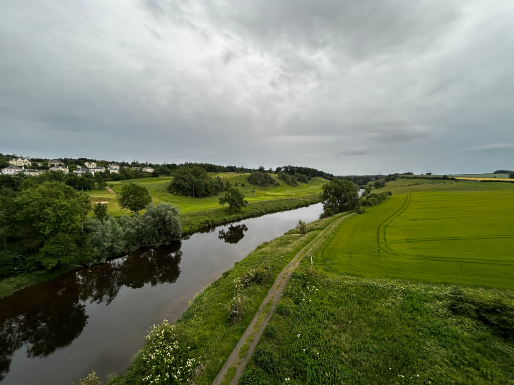 High-angle, long shot of a tranquil river meandering through a lush green landscape under a cloudy sky. On one bank, there's a golf course visible with residential buildings in the background. The other side features a sprawling field of crops. A small, unpaved path runs alongside the river.