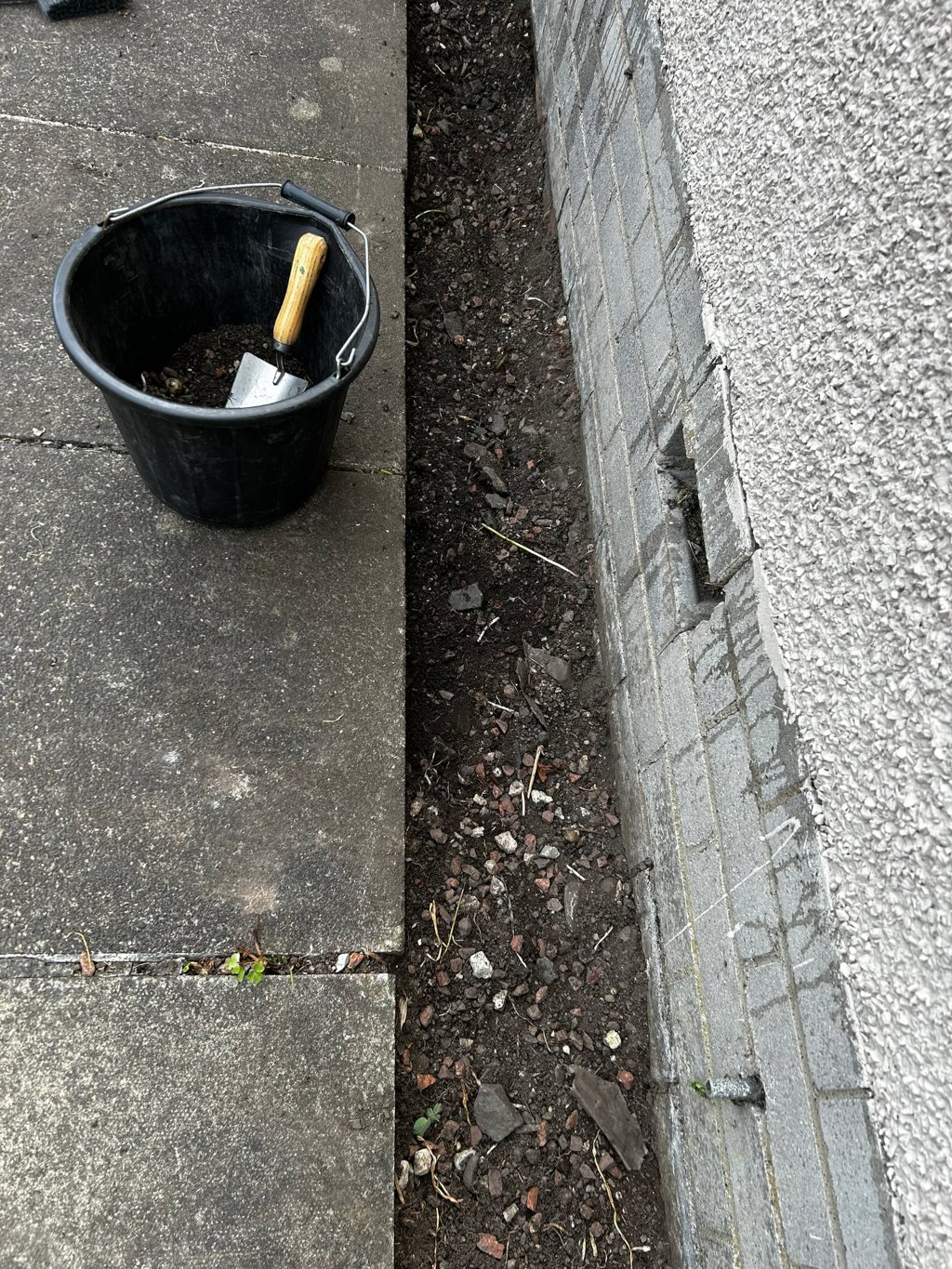 Black bucket with a small trowel inside, sitting on a concrete surface next to a narrow strip of earth bordering a grey brick wall. The earth contains small stones and debris. The scene suggests some form of minor gardening or landscaping work in progress.