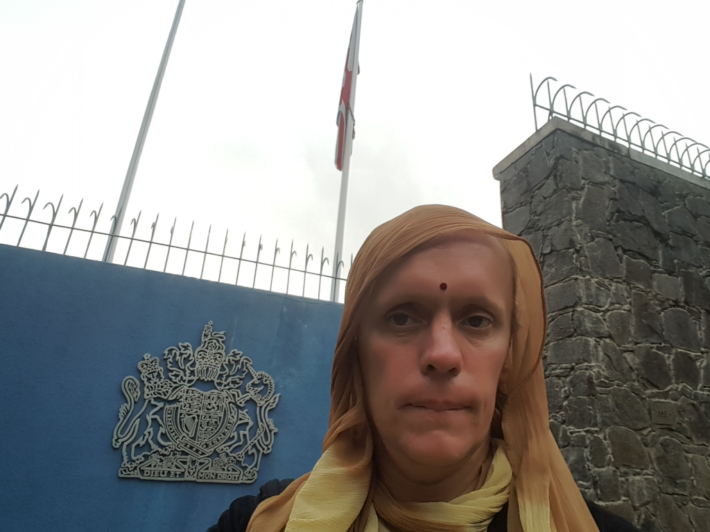 Leonie wearing a light tan, almost beige, headscarf draped loosely around their head and shoulders. The scarf is sheer enough to allow the person's face and hair to be partially visible. A small, dark red bindi is centred on their forehead between their eyebrows. Their expression is serious, almost sombre, with a neutral mouth and direct gaze toward the camera. The person appears to be standing in front of a wall. The background features a section of a light blue wall, a tall metal fence with sharp points at the top, and a stone wall that's dark gray and brown, constructed from rough-hewn stones. A tall flagpole stands behind the fence, hoisting a Union Jack flag, which is partially visible and seems to be slightly off-centre to the right. The overall lighting is diffused, indicating an overcast sky or indirect light. The colour palette is muted: the blue of the wall, the beige of the headscarf, the gray and brown of the stone, and the somewhat subdued colours of the flag. On the blue wall, there is a large, silver-gray emblem—the coat of arms of the United Kingdom—with the words DIEU ET MON DROIT visible beneath it.