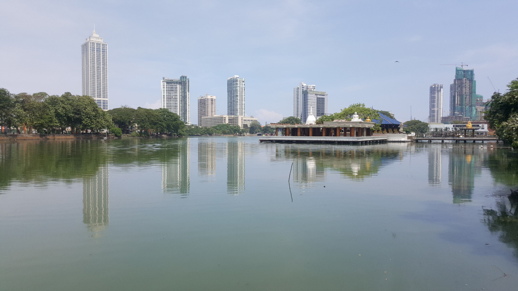 Calm, greenish-grey lake reflecting a cityscape and a modern, light-brown structure. The structure, situated centrally in the lake's middle ground, appears to be a pavilion or temple with a flat roof and distinct pillars. It has a white, slightly elevated central section. Surrounding the lake are lush, dark-green trees forming a natural border along the shoreline. In the background, several modern skyscrapers of varying heights rise against a pale, almost cloudless sky. One particularly tall, slender skyscraper dominates the left side. Another, more blocky building is under construction with a visible crane. These buildings are rendered with sharp lines and distinct architectural details. There are no people visible in the image.