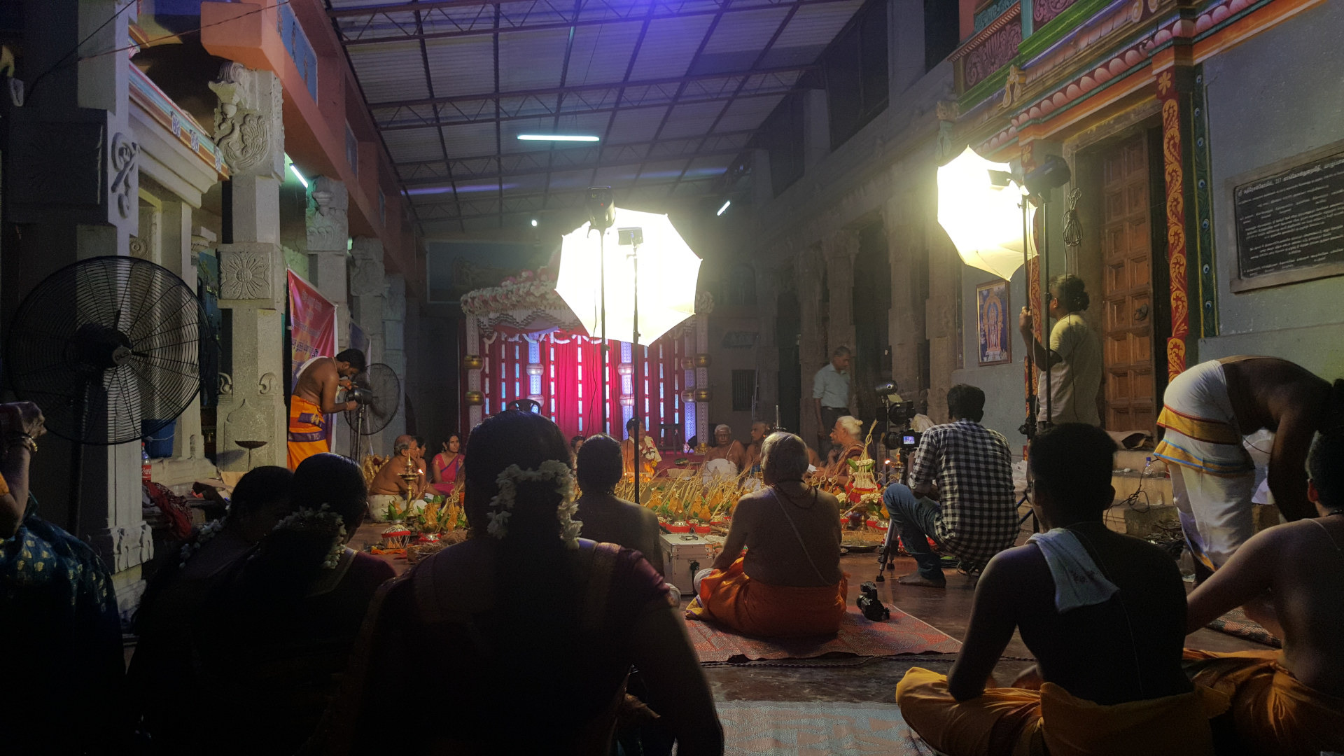 Hindu religious ceremony or ritual, possibly a puja, taking place within a temple or temple-like structure. The main focus is on a group of people seated on the floor, many dressed in traditional South Indian attire – vibrant saris and dhotis in shades of orange, yellow, and red. Some individuals wear garlands of white flowers. In the center foreground, a woman with a dark sari and a flower garland in her hair sits with her back to the viewer, her posture suggesting focused attention. Several people are positioned around the central group, including those who appear to be videographers or photographers, holding professional-looking cameras and lighting equipment (softboxes). One man, dressed in a traditional orange dhoti, is standing slightly to the left, appearing to be a participant in the ritual. Another man, wearing a plaid shirt, is kneeling, operating a camera. Other individuals seem to be assisting in the setup and filming. At the right edge, a man in a white dhoti is engaged in some task seemingly related to the ceremony.
