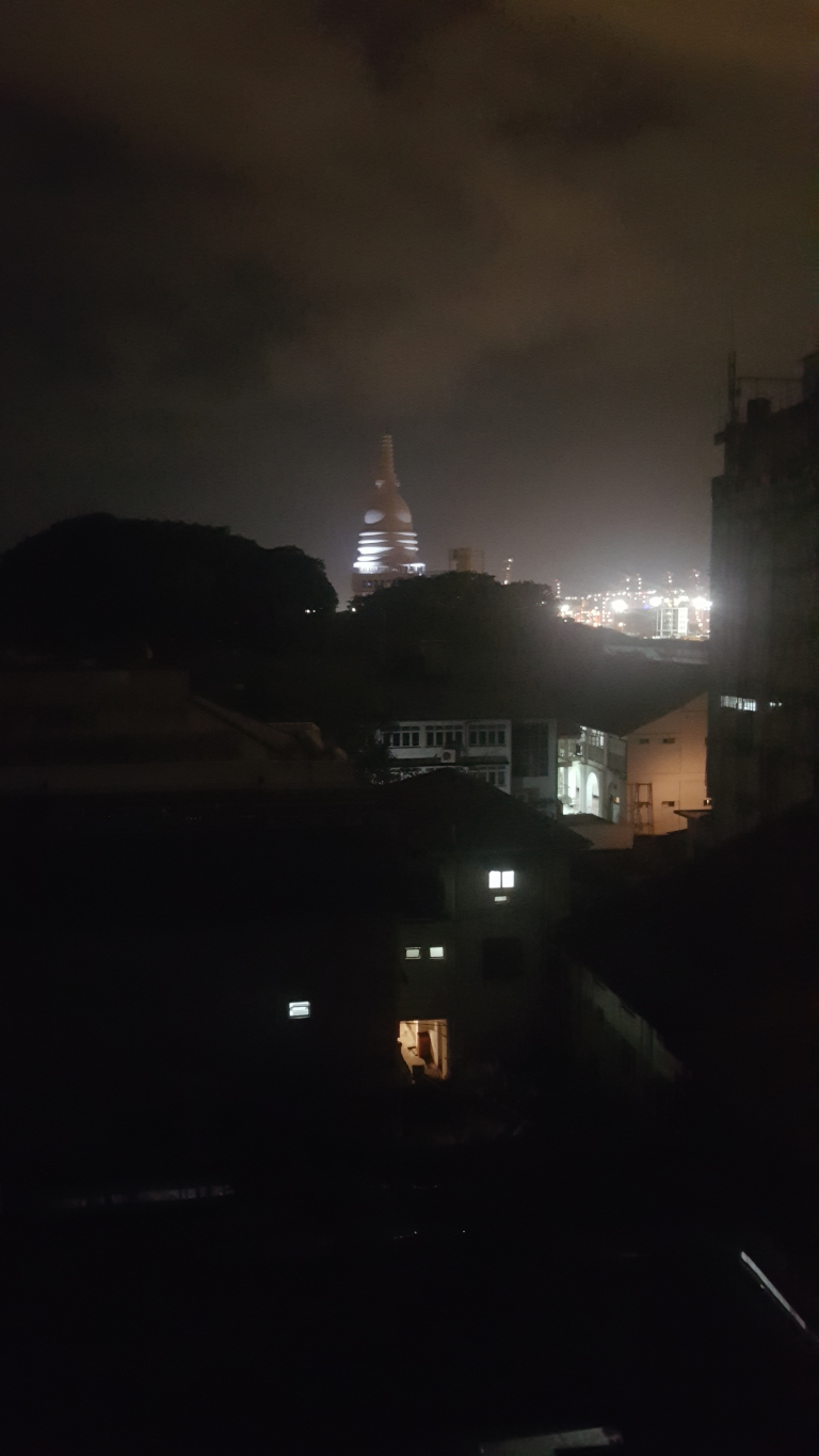 Distant, brightly lit stupa, a dome-shaped Buddhist monument, standing tall and slender against the night sky. It's centrally positioned, but slightly off-center, creating a subtle visual imbalance. In the foreground, several dark, silhouetted buildings are visible, their outlines suggesting residential structures with dimly lit windows. The buildings are rendered almost entirely in shadow, with only a few internal lights breaking through the darkness. A portion of a larger, possibly multi-story structure, is visible on the right edge of the frame, also partially obscured by darkness.