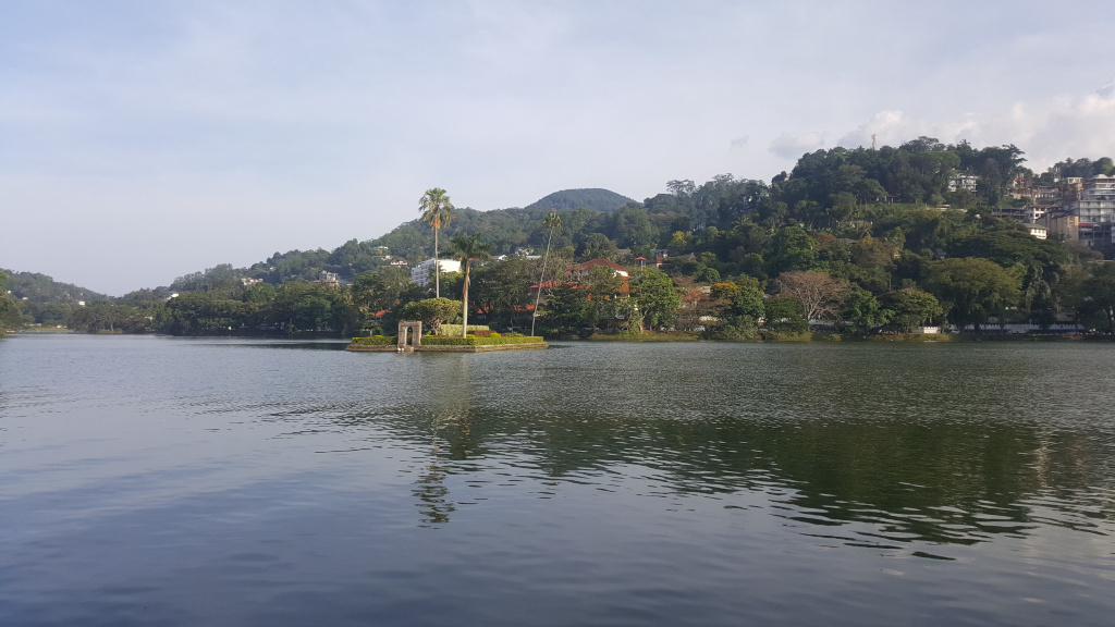 Serene lake, its dark, still water reflecting the surrounding landscape. A small, verdant island sits prominently in the mid-ground. This island features lush tropical vegetation, including several palm trees, and a small, aged-looking stone structure, possibly an arch or a small pavilion, near its centre. The structure appears to be made of light-grey or tan stone, weathered by time and exposure to the elements. There are no people visible in the image. The scene is set in a tropical or subtropical climate. The background is dominated by a gently sloping hill densely covered with lush green vegetation. Houses and buildings are interspersed within this greenery, appearing mostly as residential structures, scattered among the trees. The hills rise smoothly to meet the sky. The sky is a soft, light blue, with just a hint of cloud cover, suggesting a calm, possibly slightly overcast day. The lighting is soft and diffused, indicative of either early morning or late afternoon light. There are no harsh shadows. The overall colour palette is muted yet vibrant: dark greens of the foliage, darker greys and browns of the buildings and stone structure, the dark grey-blue of the calm lake, and the pale, washed-out blue of the sky.
