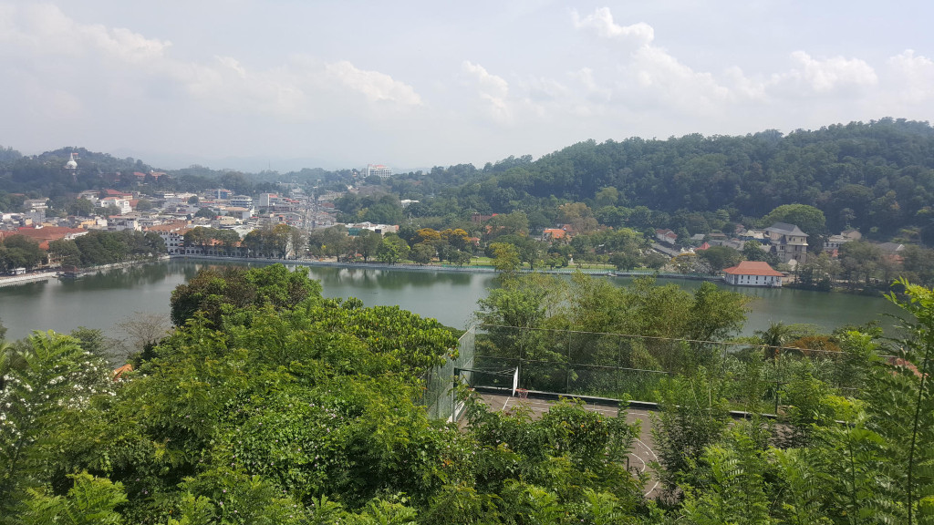 Panoramic view of Kandy Lake, Sri Lanka, nestled within a vibrant green landscape. The lake itself is a calm, dark teal, reflecting the surrounding sky and foliage. A portion of a basketball court is visible in the foreground, partially obscured by lush vegetation. The city of Kandy sprawls across the background, showcasing a mix of low-rise buildings in muted earth tones, punctuated by occasional white structures. Hills heavily forested with deep green trees rise gently beyond the lake and the city, creating a layered backdrop. A white structure, possibly a stupa or temple, is faintly visible on a distant hill to the left. There are no people visible in the image.
