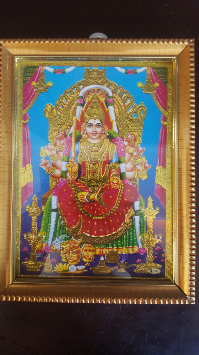 Richly adorned goddess seated majestically on a golden throne. She is the focal point, occupying most of the frame. Her attire is extravagant: a vibrant red and gold sari embellished with intricate patterns and sparkling details, suggesting opulence and divinity. Her complexion is light, and her expression is serene yet regal. She wears heavy gold jewelry, including necklaces, bangles, earrings, and a crown, all meticulously detailed. Her multiple arms (characteristic of many Hindu goddesses) are gracefully posed, holding various symbolic objects – possibly a weapon, a flower, or a blessing gesture. At the bottom of the image, three severed male heads are depicted, a common motif in some depictions of the goddess, symbolising the triumph of good over evil.