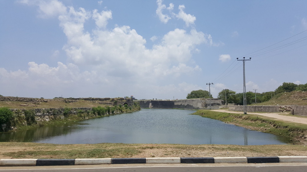 Tranquil moat, a long, narrow body of still, dark-greenish water, reflecting the sky and the surrounding structures. The moat is bordered by low, grassy banks and runs parallel to a section of ancient, weathered stone walls, which form a low, fortress-like structure. These walls, a muted gray-brown, show signs of age and wear, with patches of vegetation sprouting from cracks and crevices. Beyond the wall, a flat, grassy expanse extends to the horizon. Power lines stretch across the right side of the image, leading the eye towards a slightly elevated area with sparse, low-lying trees. The foreground features a simple, black-and-white painted curb along a paved road, separating the viewer from the moat. No people or animals are visible.