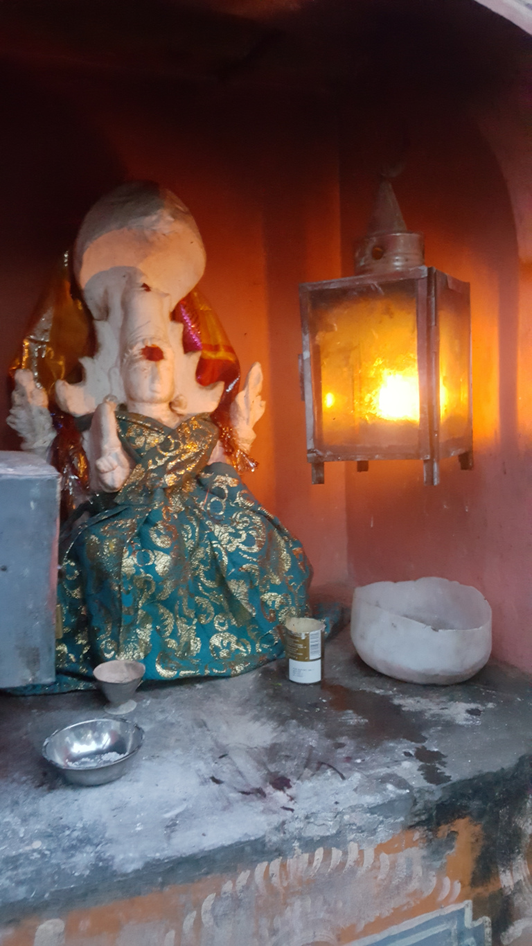 Small, intricately dressed statue of what appears to be a Hindu deity, possibly a form of Ganesha. The statue is predominantly white or light beige, with a rounded head and what seems like elephantine features, though partially obscured. It’s adorned in a vibrant teal and gold patterned garment, draped in flowing folds. The dress is richly decorated with gold accents, giving a sense of opulence. The statue's hands are posed in a traditional devotional gesture. To the right, a simple, hanging lantern made of glass and metal casts a warm, orange glow. Inside, a small flame burns brightly, illuminating the scene. Near the base of the statue, there are small bowls, one metallic and possibly containing ash or some other offering, and another white bowl which could be used for offerings. A small, cylindrical container, possibly a small tin or candle, sits near the lantern.