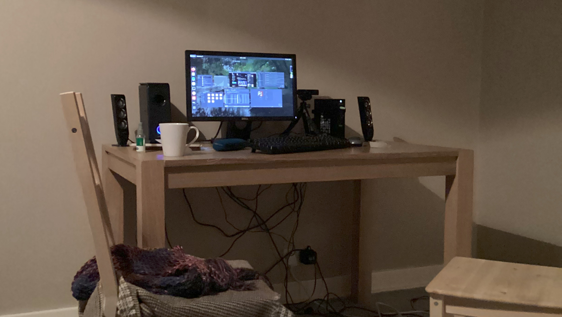 Simple, lightly coloured wooden desk in a dimly lit room. On the desk is a computer monitor displaying multiple windows, a keyboard, a webcam on a tripod, computer speakers, and a mug. There are also what appear to be some other electronic devices and a tangle of wires under the desk. A wooden chair with a knitted throw is partially visible to the left. The overall impression is of a home workspace or gaming setup in a subdued, perhaps late-evening or nighttime, setting.