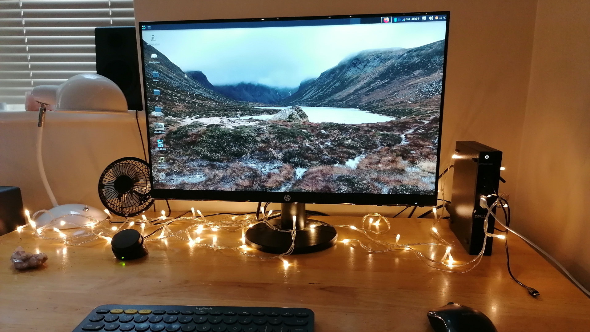 Computer desk at night, illuminated by warm-toned fairy lights. The centrepiece is an HP monitor displaying a serene landscape wallpaper of a valley with a lake. A small computer tower and other peripherals (keyboard, mouse, fan) are visible. The overall mood is calm and cosy, suggesting a personal workspace. The fairy lights add a festive or homely touch.