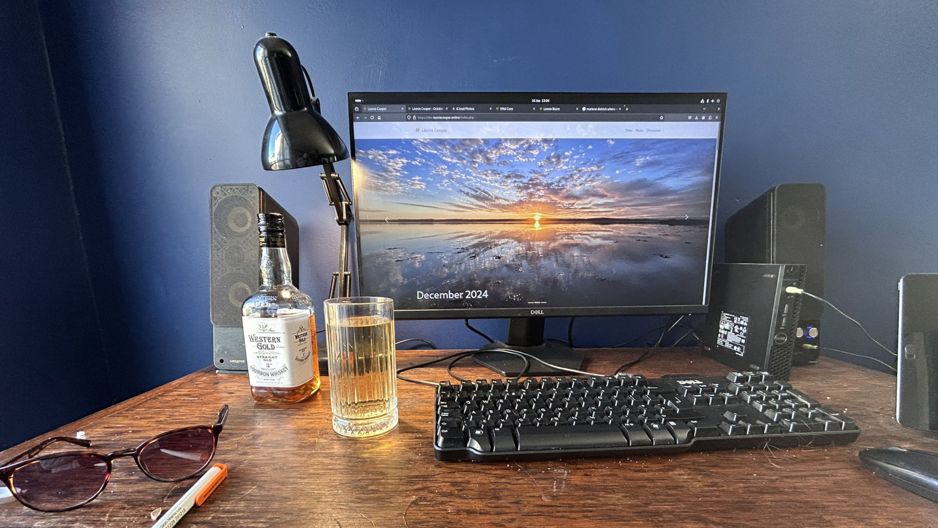 Desk setup with a computer monitor displaying a panoramic sunset over a calm body of water. The date “December 2024” is visible on the screen. On the desk are various items including a bottle of Western Gold bourbon whisky, a glass of what appears to be a light-coloured alcoholic beverage, a keyboard, computer speakers, a desk lamp, a pair of sunglasses, and a pen. The overall aesthetic suggests a calm, possibly contemplative atmosphere.